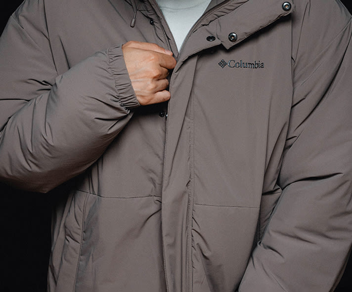 Person wearing a taupe Columbia insulated jacket with hood and black bucket hat against a dark background, adjusting the front zipper.