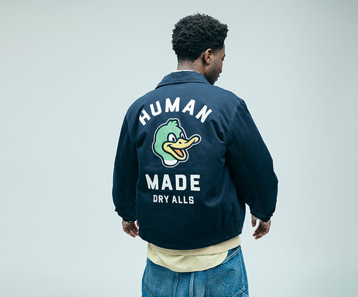 Back view of a person wearing a navy blue HUMAN MADE jacket with a large duck graphic and white “HUMAN MADE” and “DRY ALLS” lettering on the back, paired with blue denim jeans against a light grey background.