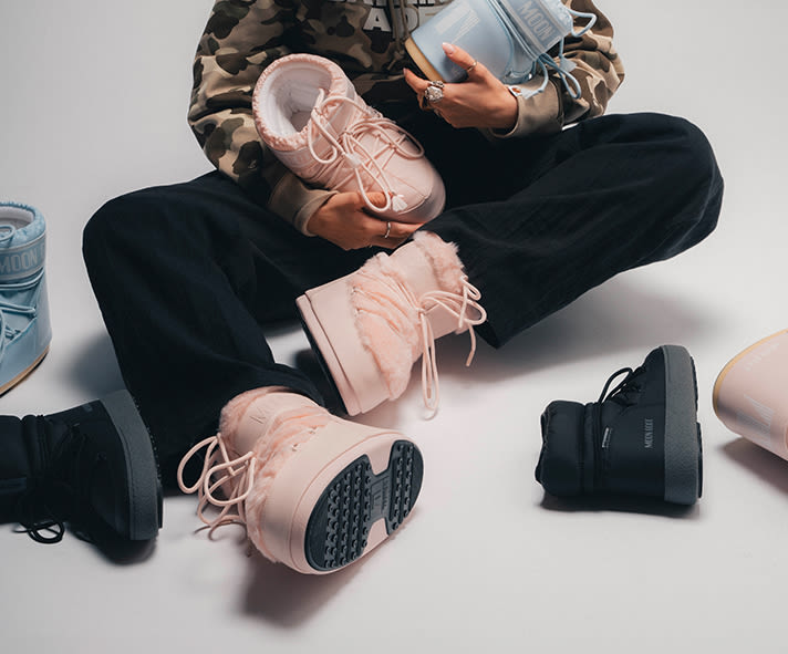 Person seated on the floor wearing light pink Moon Boot winter boots, holding a light blue Moon Boot model while dressed in a camouflage-print hoodie and dark trousers.