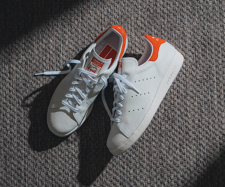 Pair of adidas Stan Smith 80s (Cracked Leather) sneakers in white with orange heel tabs, displayed on a textured carpet, highlighting the cracked leather upper and classic tennis-inspired silhouette.