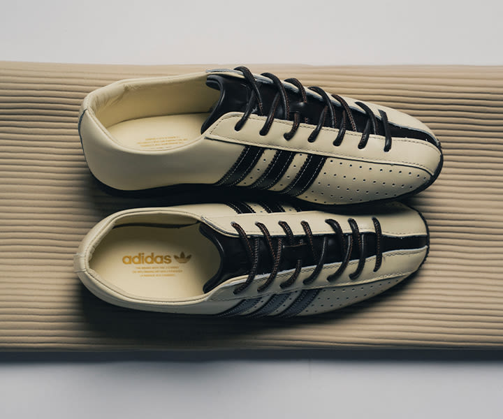 Top view of adidas Japan Lux sneakers in off-white leather with black three stripes and dark laces, displayed on a ribbed beige surface, highlighting the premium materials and streamlined vintage silhouette.