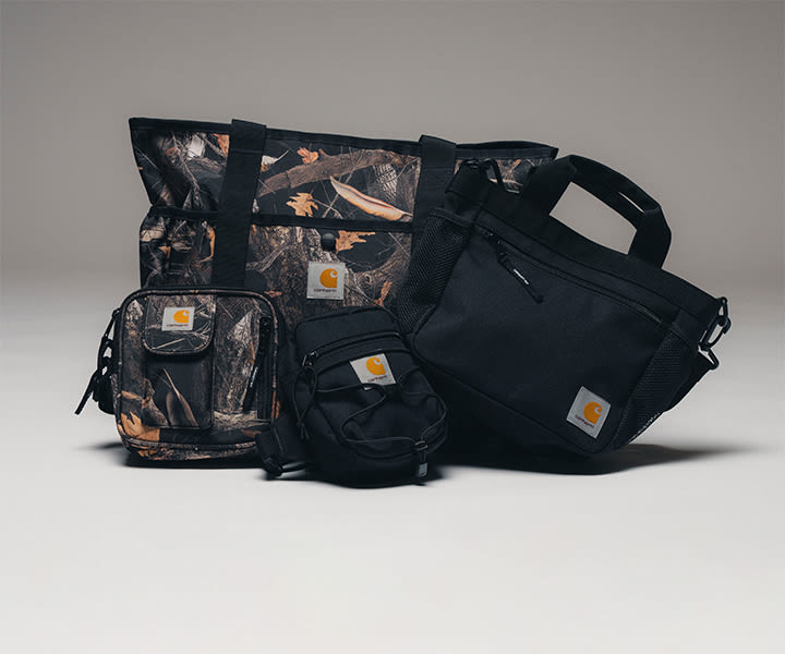 Selection of Carhartt WIP bags, including black and camouflage shoulder bags, a utility pouch, and a tote bag, displayed together against a clean studio background.