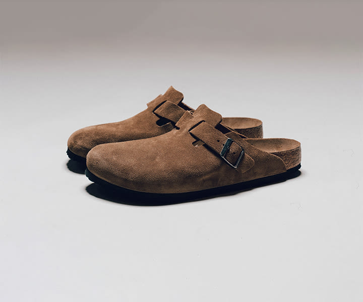 A pair of brown suede Birkenstock Boston clogs, featuring a single strap with a silver buckle, resting side-by-side on a light-colored surface.