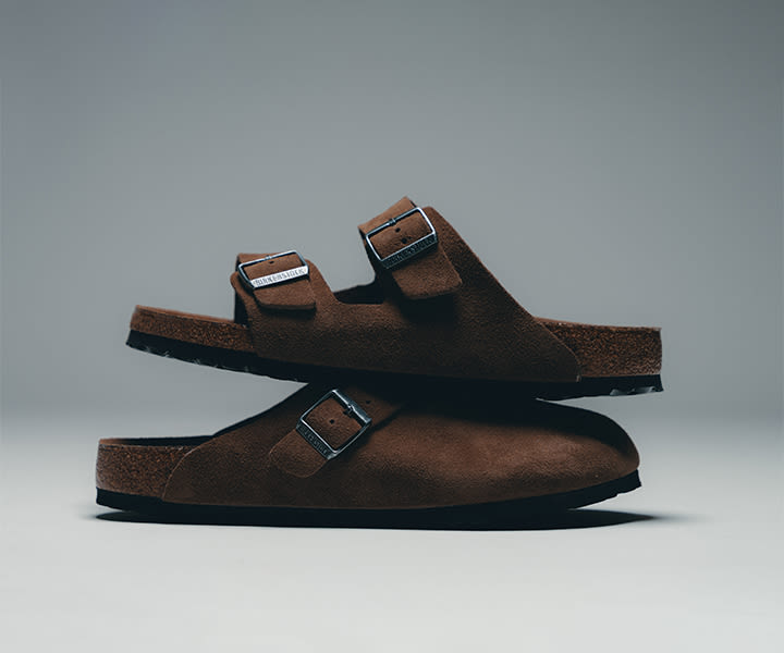 A pair of brown suede Birkenstock sandals with two adjustable straps and silver buckles, stacked one on top of the other against a neutral grey background.