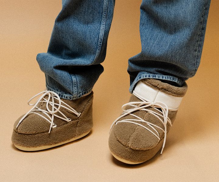 The image features a close-up of a person’s feet wearing plush, cozy moon boots. The boots are brown, with a soft, fleece-like material and white laces, paired with casual blue jeans. The light background highlights the cozy and relaxed vibe of the footwear. The boots appear to be designed for comfort and warmth.