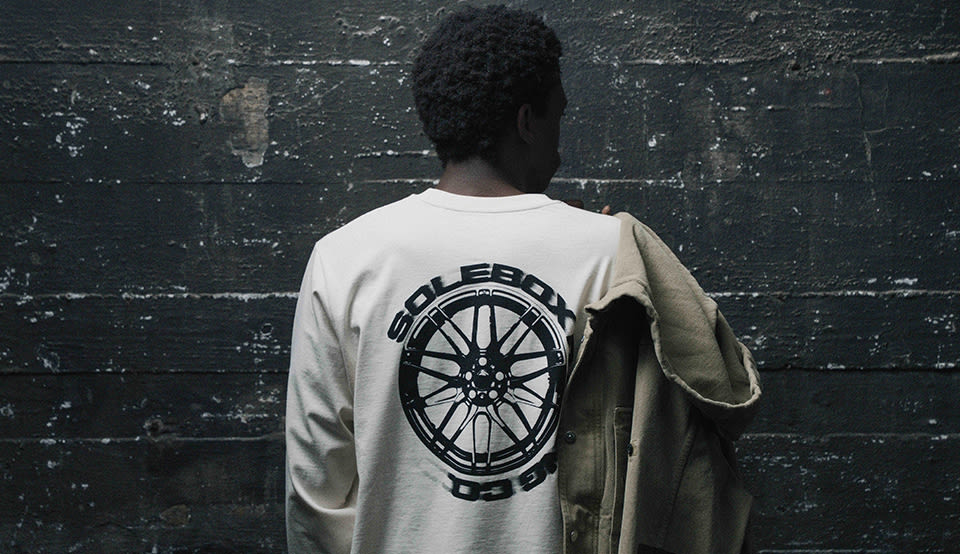Person seen from behind wearing a cream long-sleeve shirt with a large solebox wheel graphic on the back, holding a beige jacket over one shoulder against a dark industrial wall.
