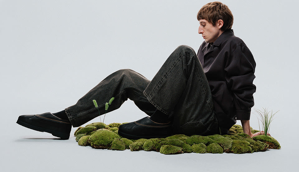 Person seated on a moss-covered patch in a minimalist studio setting, wearing dark denim and a dark jacket with black SOREL slip-on shoes featuring waterproof rubber uppers and suede detailing. The neutral backdrop and natural elements emphasize the outdoor-inspired design and everyday versatility of the footwear. Geben Sie Ihr Feedback zu BizChat ab.