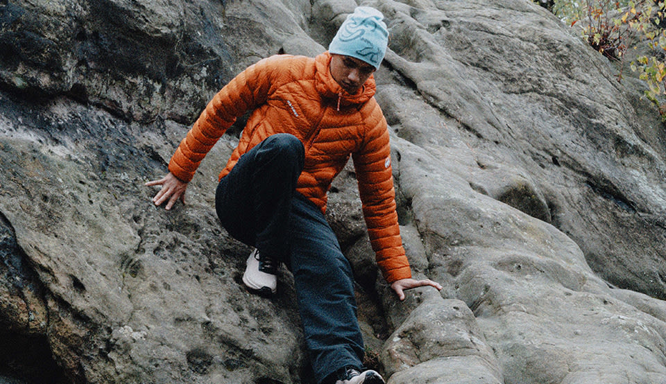Person wearing an orange Mammut insulated hooded jacket while standing in front of a rugged rock face outdoors.
