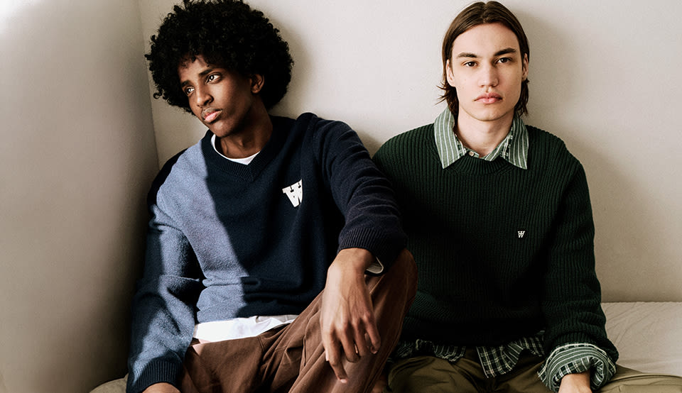 Two models wearing Wood Wood knitwear and layered casual outfits, seated indoors against a neutral wall in a minimalist setting.