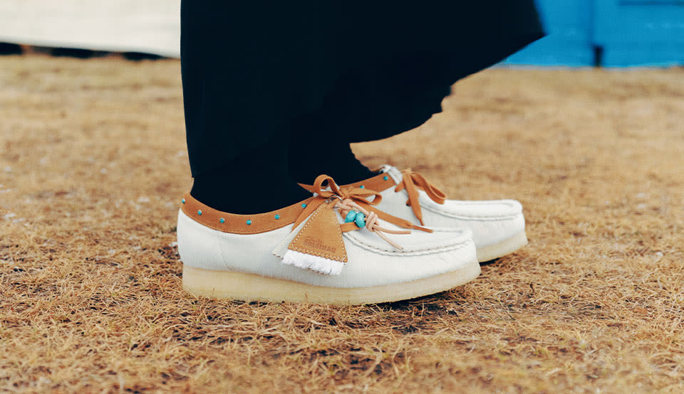 Close-up of Clarks Wallabee shoes in white textured suede with tan leather detailing, decorative tassels and signature crepe soles, worn outdoors on dry grass.