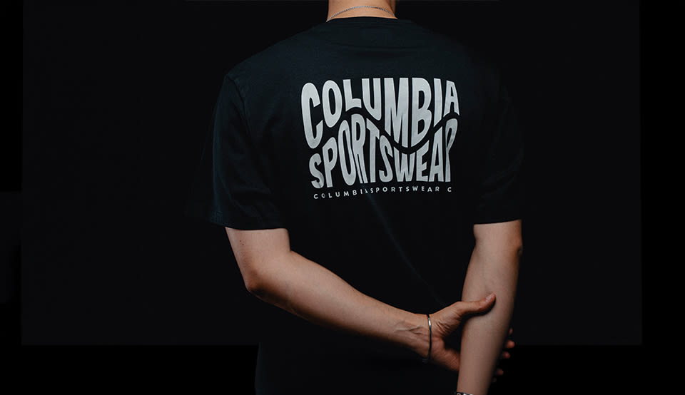 Alt Text Image Creator said: Alt Text Image Creator Alt Text (EN): Back view of a person wearing a black Columbia Sportswear T-shirt with a large white Columbia Sportswear logo print on the back against a dark background.
