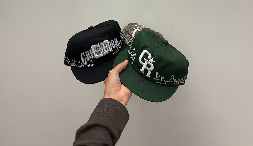 Hand holding two embroidered snapback caps in black and dark green with large “CR” logo and horse stitching details on a plain background.