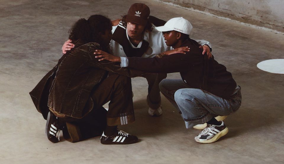 Three models crouching in a circle wearing pieces from the adidas x SONG FOR THE MUTE collection, styled in earthy tones with adidas footwear and apparel in an industrial setting.