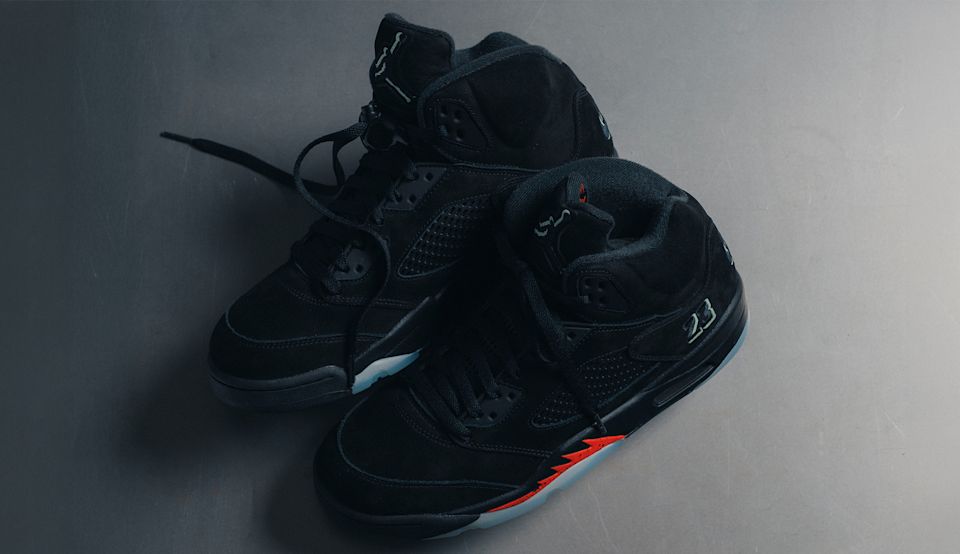 Air Jordan 5 Retro "Halloween" sneakers in black with reflective details and bright orange accents, photographed on a metallic surface.