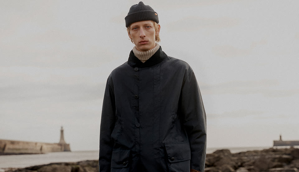 A person standing on a coastal path wearing a navy waxed jacket, dark trousers, a ribbed beanie, and suede shoes, holding a brown Barbour tartan tote bag. Rocky shoreline and calm sea in the background under an overcast sky.
