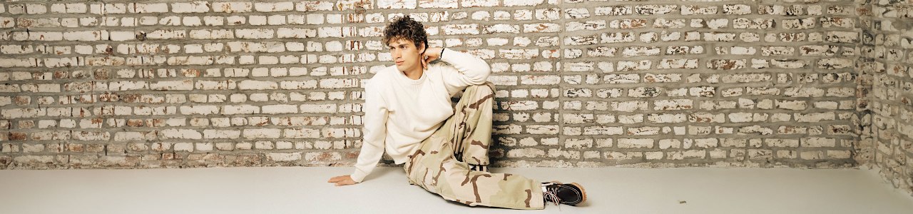 A person is sitting on the floor against a white and gray brick wall. They are wearing a loose, light-colored sweater and camouflage-patterned cargo pants paired with dark shoes. One arm rests on the floor while the other is bent, with the hand near the head. The setting has a minimalist, industrial feel with a clean, light-colored floor and textured brick background.