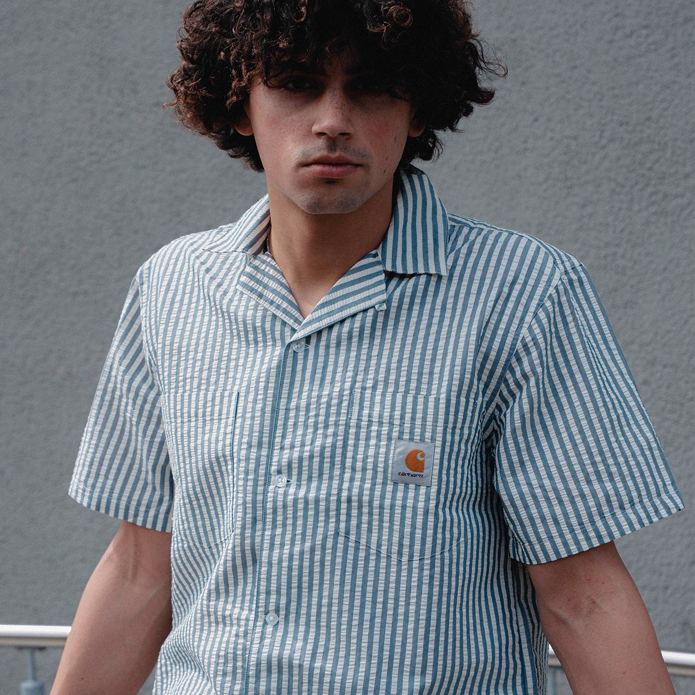 Close-up of a person with curly hair wearing a short-sleeve, blue-and-white vertically striped button-up shirt with a chest pocket and a small Carhartt logo patch, standing against a plain grey wall.