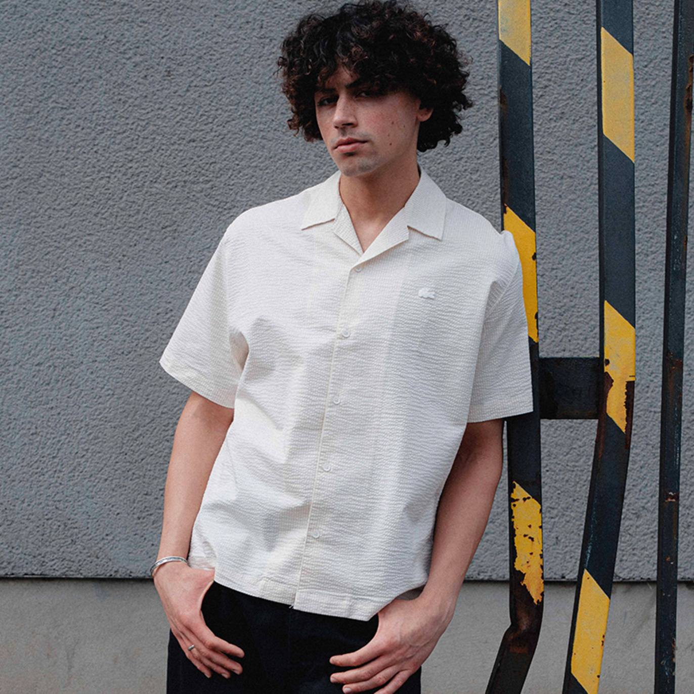Person leaning against a metal structure wearing a white short-sleeve textured button-up shirt with a small Lacoste crocodile logo on the chest, set against a grey wall with yellow-and-black striped bars.