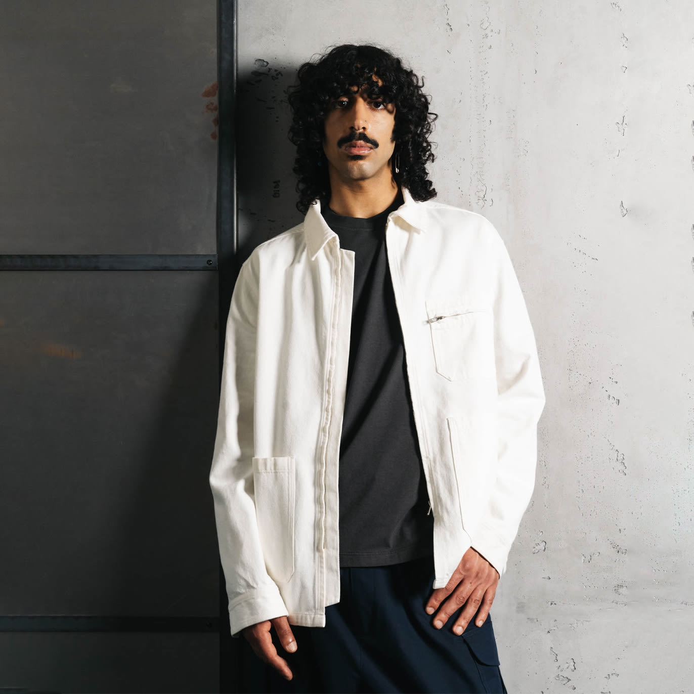 A man with curly hair and a mustache wearing a white overshirt over a black t-shirt and dark pants, leaning casually against a concrete wall with an industrial aesthetic.