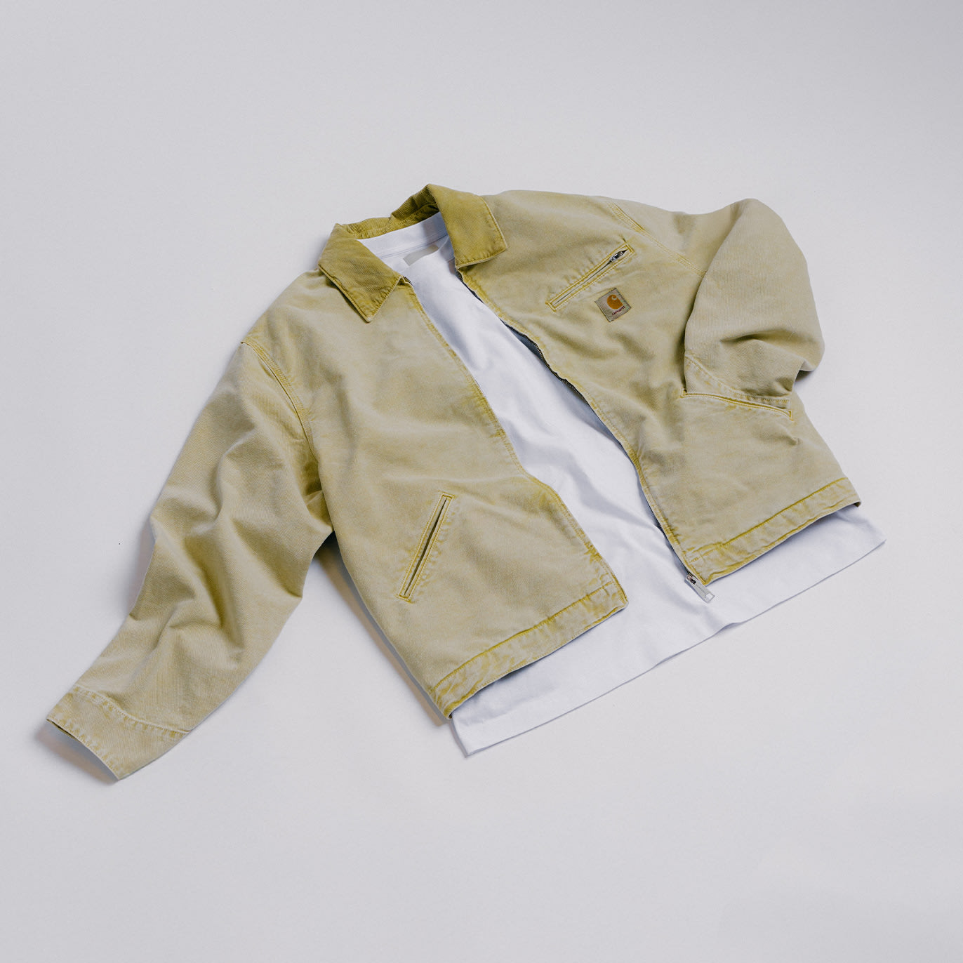 Beige workwear-style jacket laid flat over a white t-shirt on a light background.