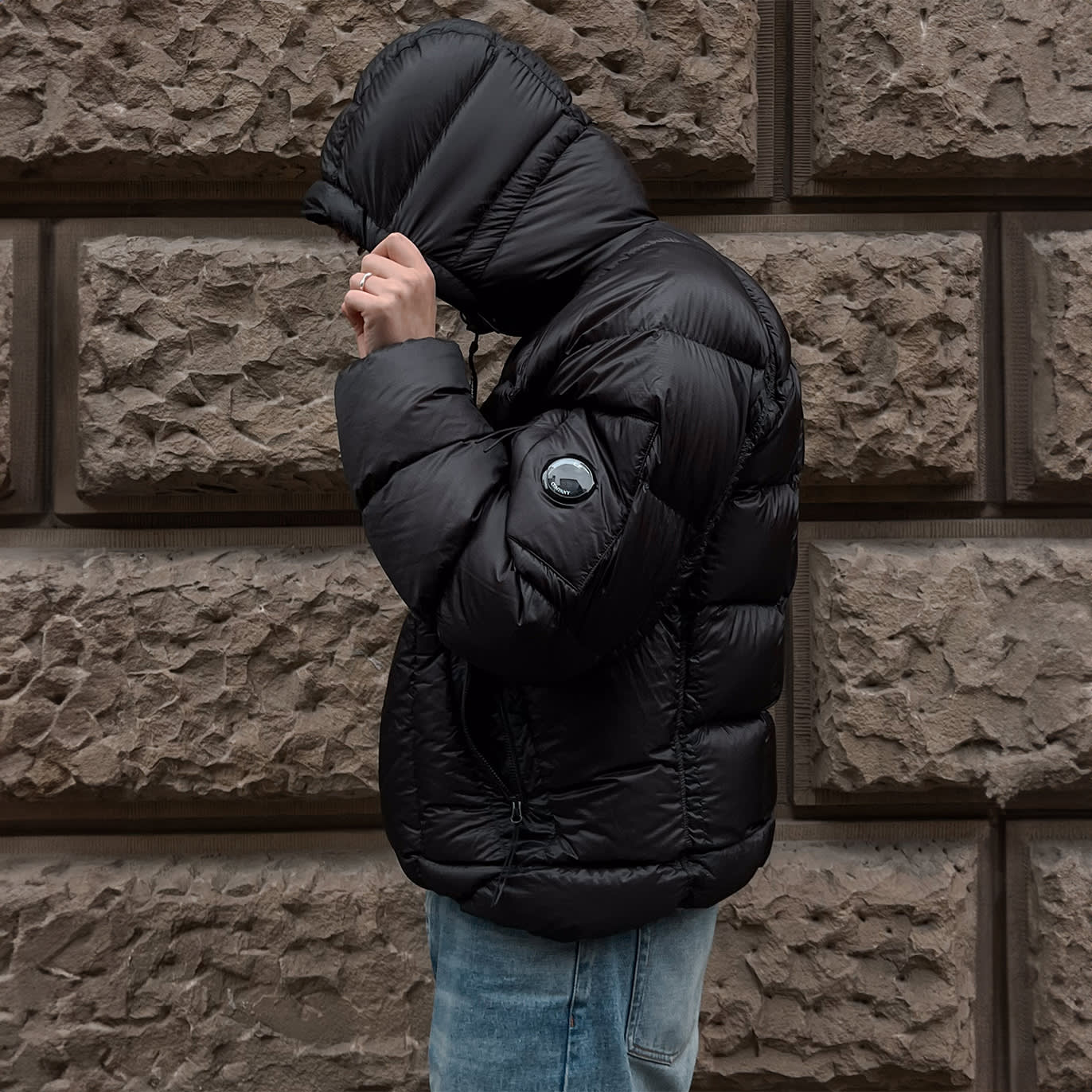 Person wearing a shiny black puffer jacket with a hood up, standing sideways against a textured stone wall.