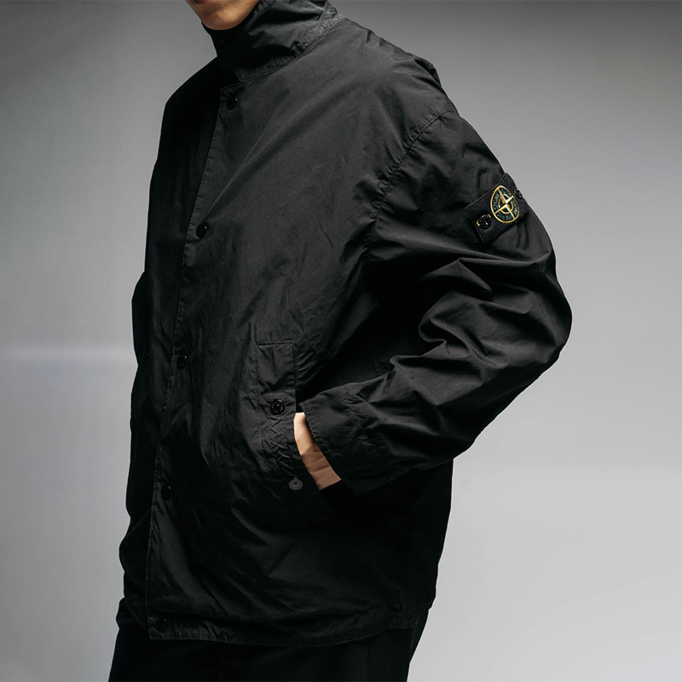 Person wearing a black lightweight jacket with a high collar and a compass patch on the sleeve, photographed against a neutral studio background.