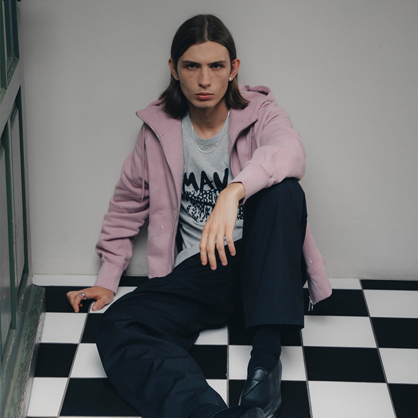 Person seated on a black-and-white checkered floor wearing a light pink zip-up hoodie over a graphic T-shirt, paired with dark wide-leg trousers and black leather shoes.