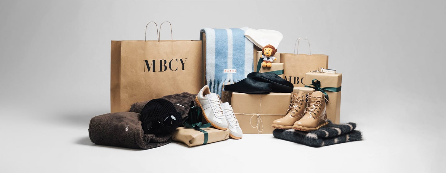 Gift arrangement with MBCY shopping bags, beige sneakers, tan boots, folded scarves, a blue striped blanket, a black beanie, and a small plush lion, all displayed on a neutral background.
