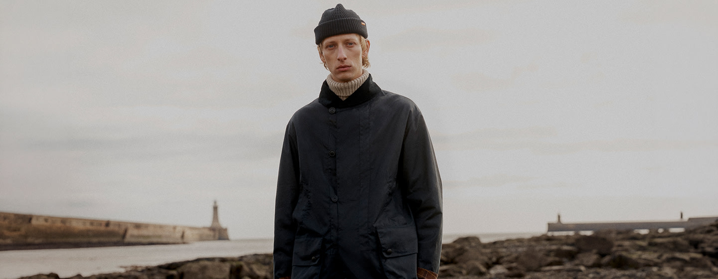 A person standing on a coastal path wearing a navy waxed jacket, dark trousers, a ribbed beanie, and suede shoes, holding a brown Barbour tartan tote bag. Rocky shoreline and calm sea in the background under an overcast sky.