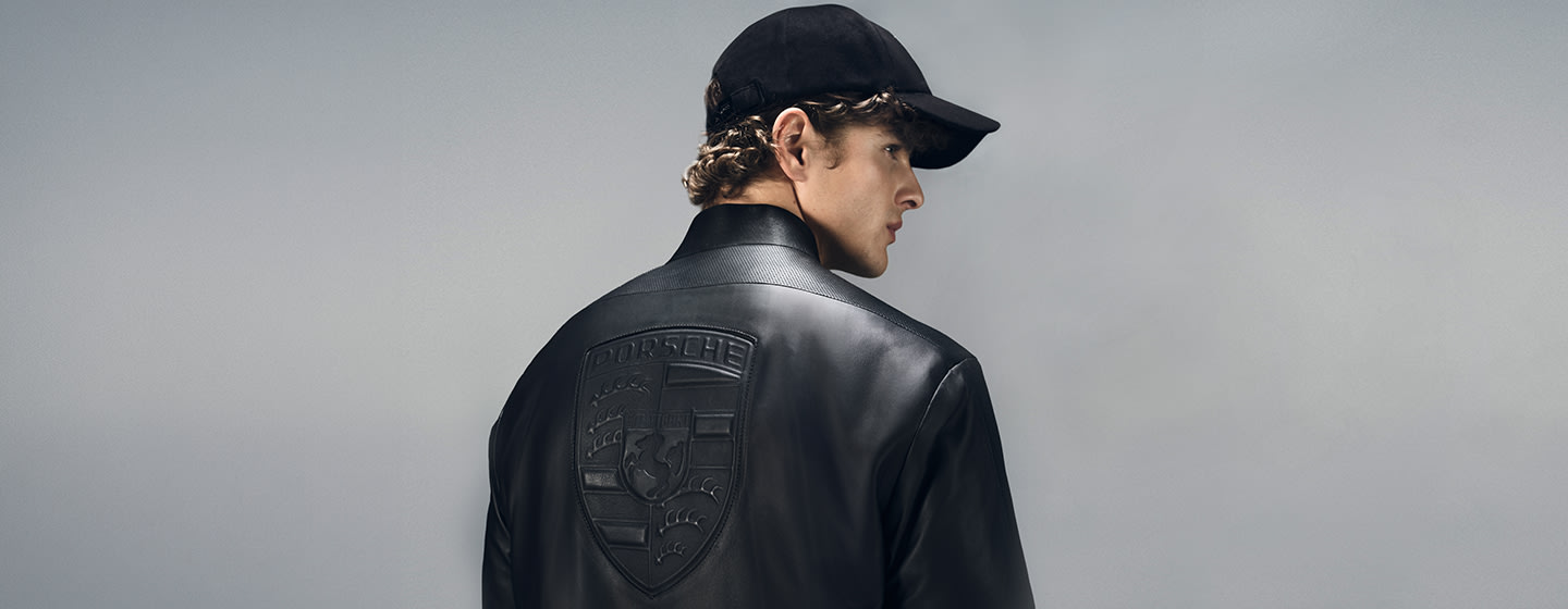 Person wearing a black BOSS x Porsche leather jacket and black cap, shown from the back with an embossed Porsche crest.