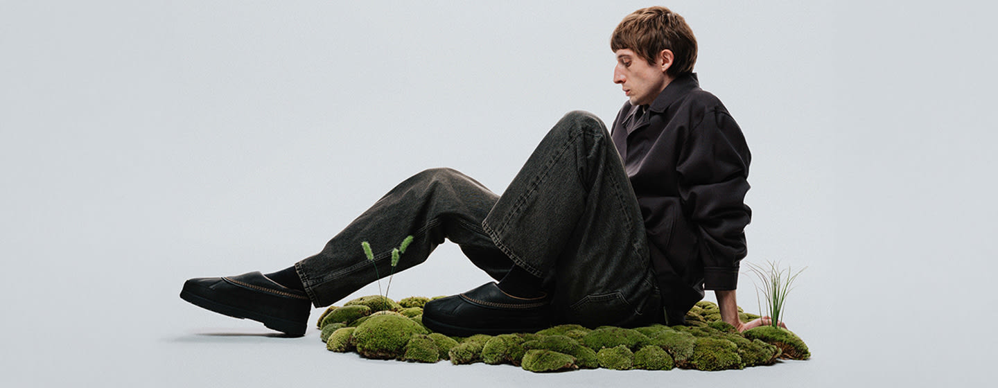 Person seated on a moss-covered patch in a minimalist studio setting, wearing dark denim and a dark jacket with black SOREL slip-on shoes featuring waterproof rubber uppers and suede detailing. The neutral backdrop and natural elements emphasize the outdoor-inspired design and everyday versatility of the footwear.