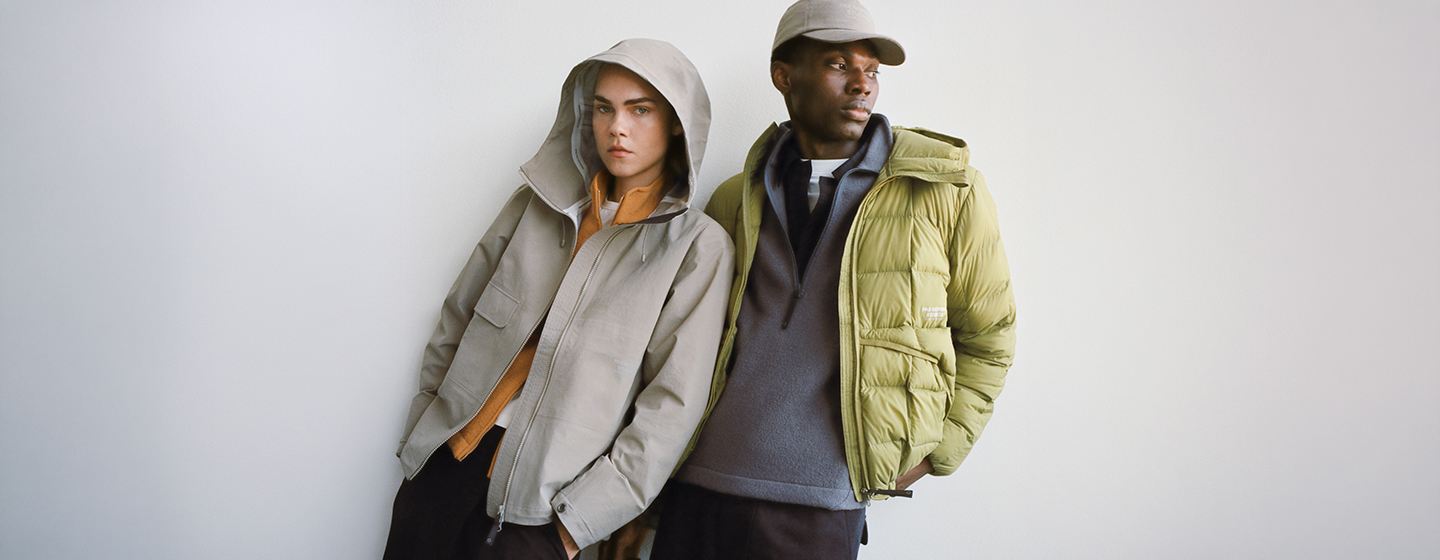 Two models wearing layered Pas Normal Studios outerwear, including a light shell jacket and an insulated puffer jacket, styled for cool-weather cycling and urban exploration.