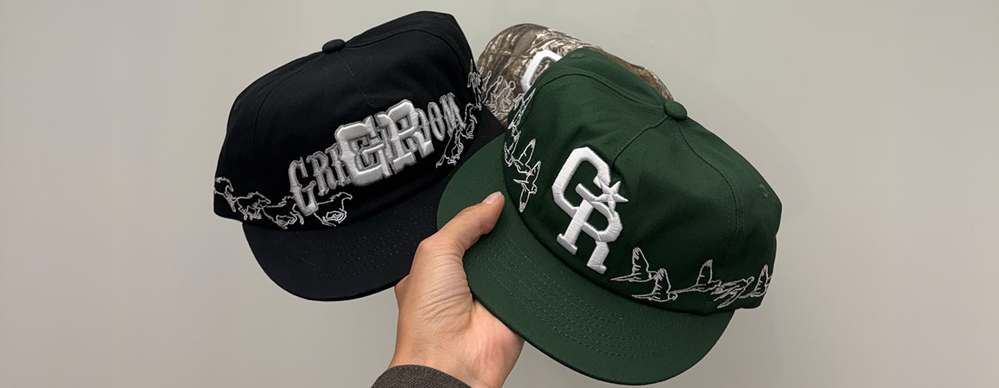 Hand holding two embroidered snapback caps in black and dark green with large “CR” logo and horse stitching details on a plain background.