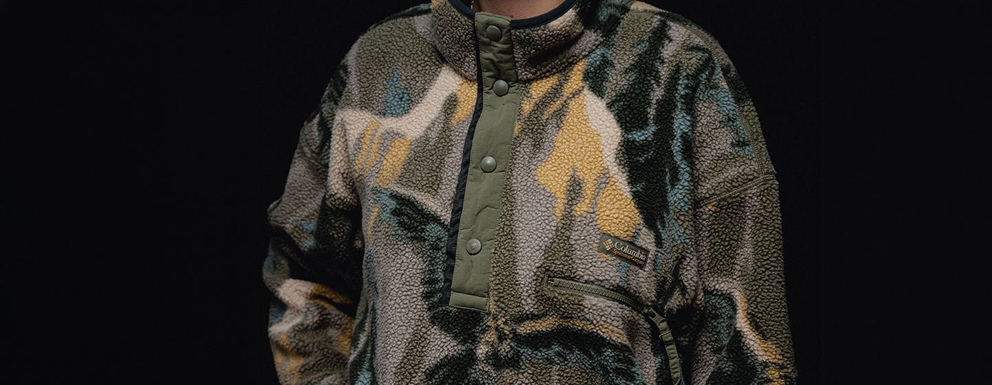 Alt Text (EN):  Close-up of a Columbia fleece pullover with an abstract mountain-inspired print in grey, green, blue and beige, featuring a snap-button collar and zip chest pocket.