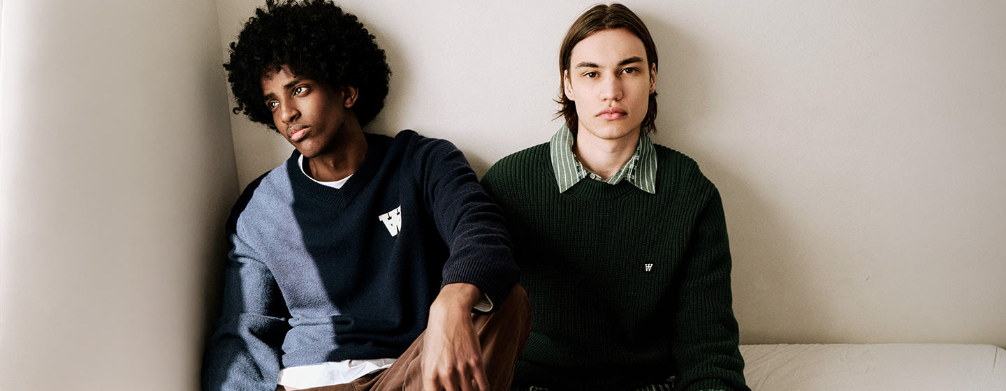 Two models wearing Wood Wood knitwear and layered casual outfits, seated indoors against a neutral wall in a minimalist setting.