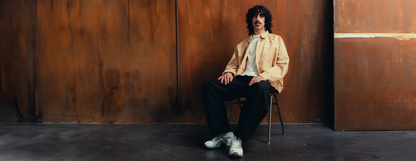 A man with curly hair and a mustache sits casually on a chair against a rust-colored wall. He wears a beige jacket over a white shirt, black pants, and white sneakers.