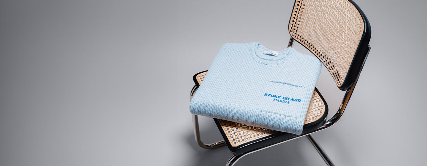 A neatly folded light blue knitted sweater with "Stone Island Marina" embroidered in blue, placed on a modern cane chair with chrome legs, set against a neutral background.