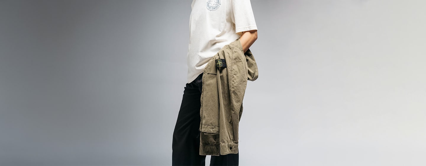 Person wearing a white short‑sleeve T‑shirt and black trousers, holding an olive‑green jacket with a compass patch on the sleeve against a neutral studio background.