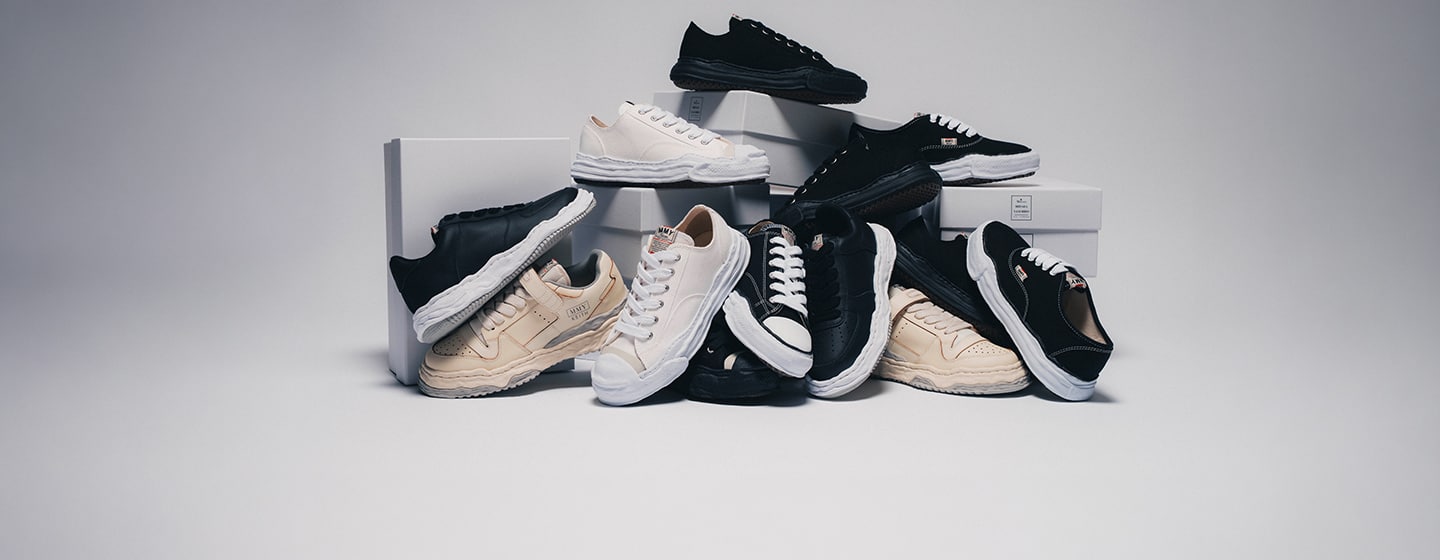 Selection of Maison Mihara Yasuhiro sneakers in black, white, and cream colorways, displayed on stacked boxes, highlighting the brand’s signature distorted soles and handcrafted-inspired design.