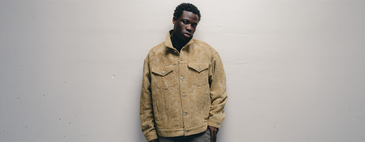 Model wearing a beige patterned Junya Watanabe MAN jacket featuring a tonal floral jacquard, button closure, pointed collar, and flap chest pockets.