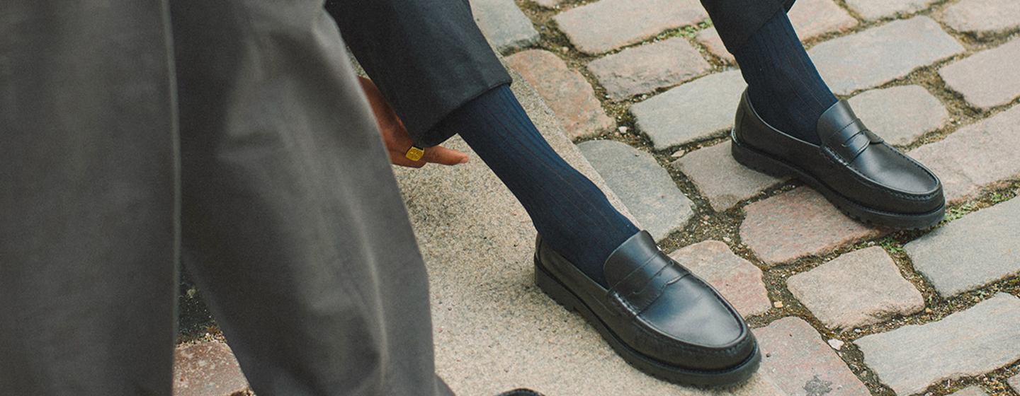 Person wearing black Vinnys leather loafers with dark socks and relaxed tailoring, seated on a cobblestone street, showcasing the brand’s refined loafer silhouette and minimalist design.  Geben Sie Ihr Feedback zu BizChat ab.