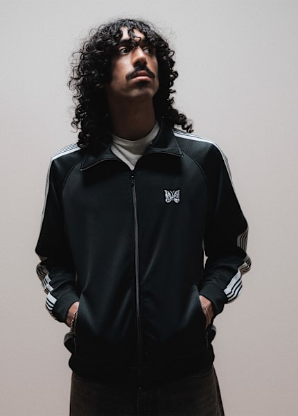 Person wearing a black Needles track jacket with striped sleeves and butterfly logo in a studio setting