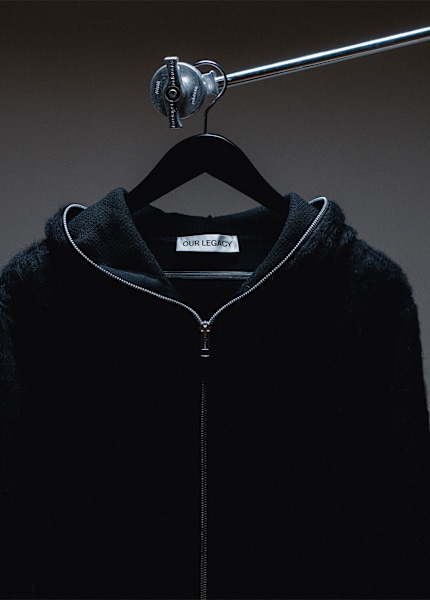 Black zip-up hoodie from Our Legacy on a hanger, displayed against a dark background.”
