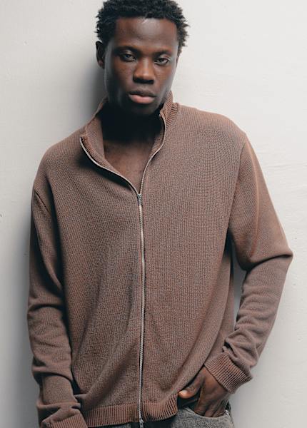 Person standing against a light-colored wall wearing an Our Legacy brown zip-up knit cardigan with a high collar, paired with relaxed-fit gray trousers, in a minimal studio setting with soft, neutral lighting.