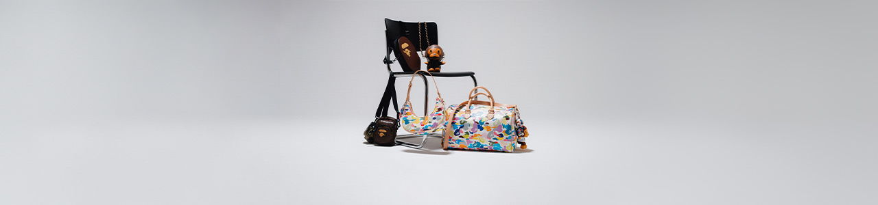 Collection of colorful BAPE bags and accessories displayed on a chair, including patterned duffle bags, crossbody bag, and monkey keychains, against a minimal light background.