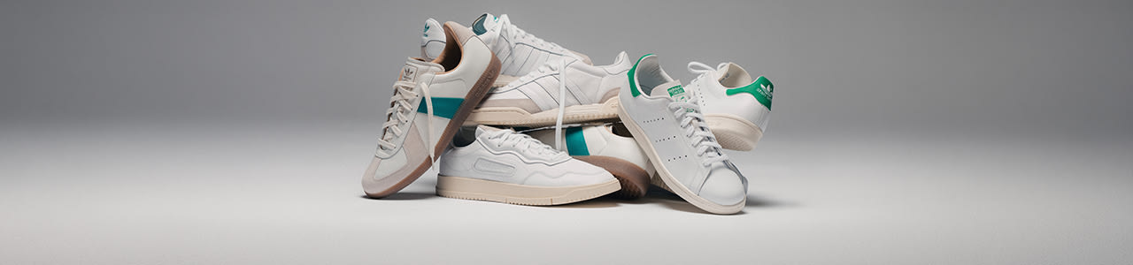 Group of adidas “House of Court Pack” sneakers stacked together on a neutral grey studio background, featuring clean white leather uppers with subtle beige and green accents, classic three-stripes branding, and gum or off-white soles.