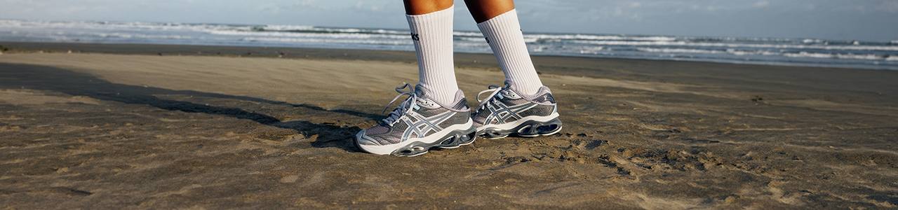 ASICS SportStyle GEL‑KINETIC 2.0 sneakers worn on sandy beach, side view of both shoes with visible cushioning sole and ASICS logo, paired with white crew socks and ocean waves in the background.