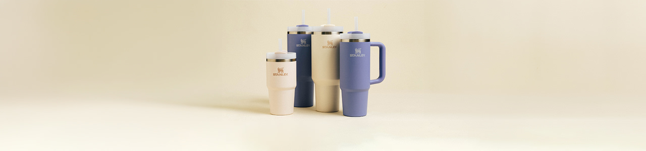 The image features a collection of Stanley brand insulated drinkware, including tumblers and mugs, in soft pastel colors (cream and lavender). The Stanley logo, featuring a winged lion, is prominently displayed on both the left side of the image and on each drinkware item. The background is a simple, warm beige color that complements the products, giving the image a clean and modern aesthetic.