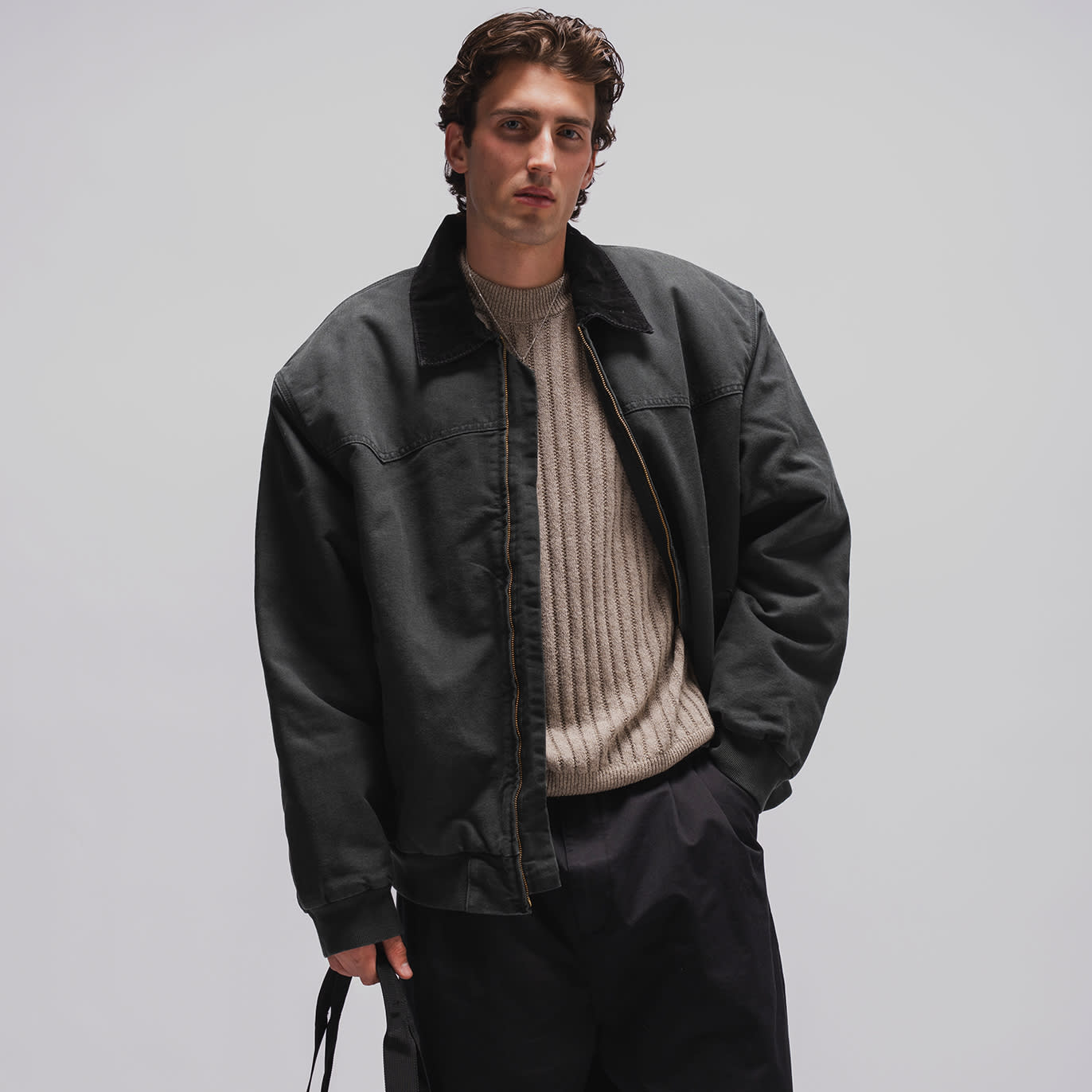 Person wearing a long dark overcoat with oversized pockets, styled with relaxed trousers in a minimalist studio setting.