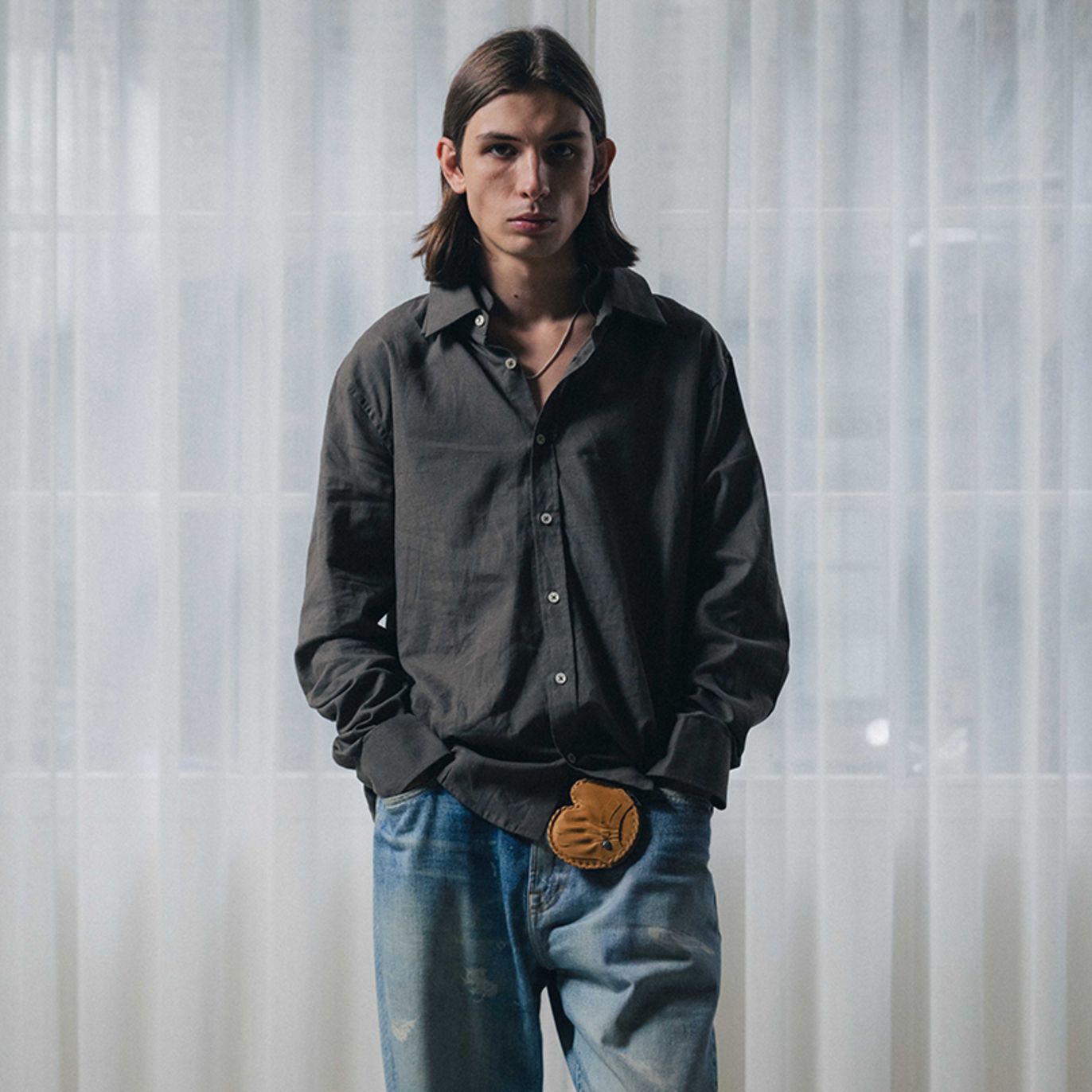 Mid-length view of a person wearing a dark button-up shirt paired with light blue washed jeans, styled with a small brown pouch attached at the waist, standing in front of sheer white curtains with hands in pockets.
